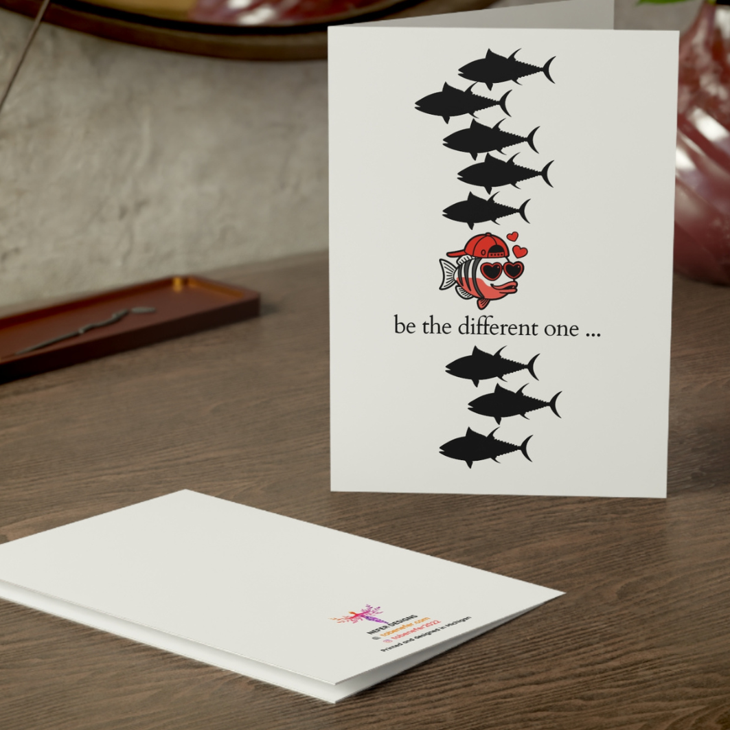 Greeting card with a unique design featuring a fish wearing sunglasses and the text 'be the different one' on a wooden surface.