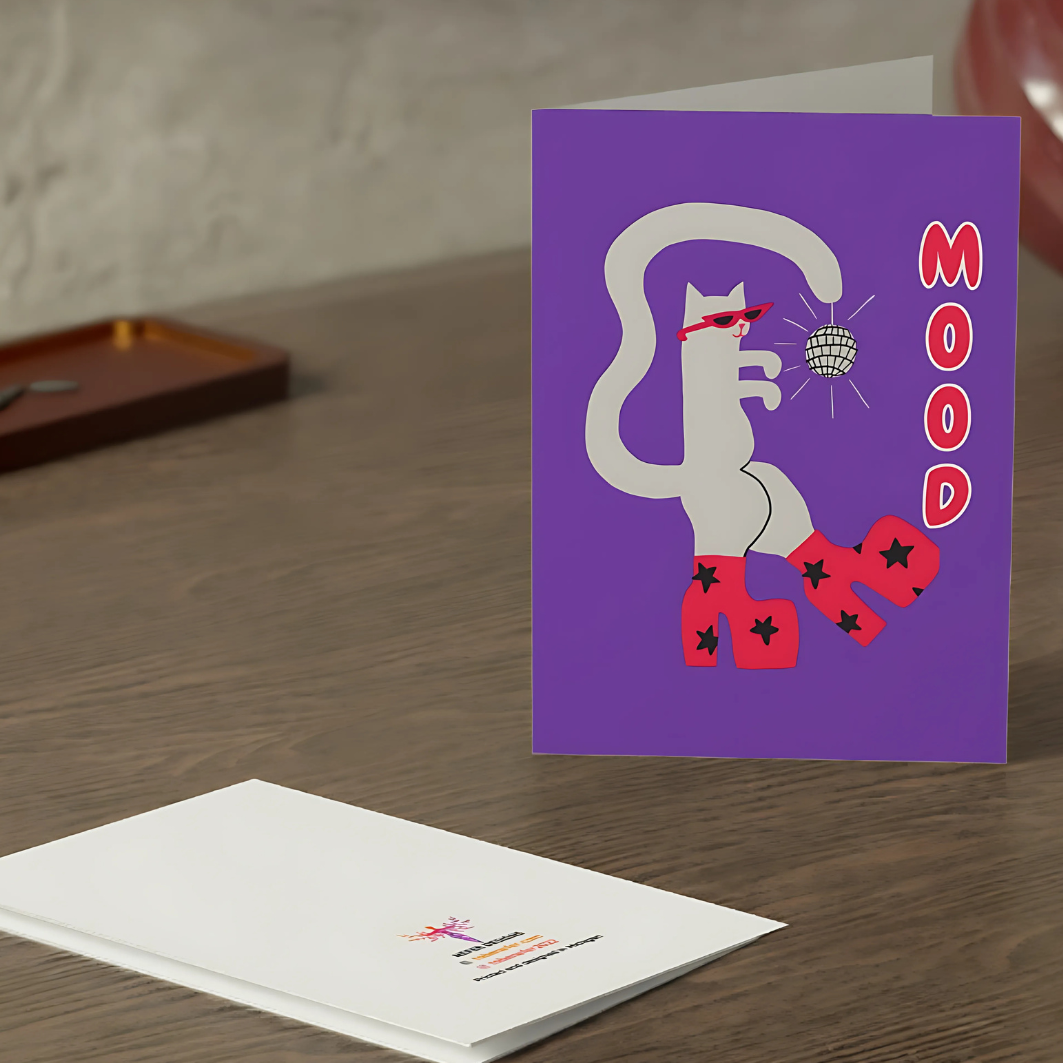Greeting card with a cat illustration and 'Mood' text on a purple background, placed on a wooden surface.