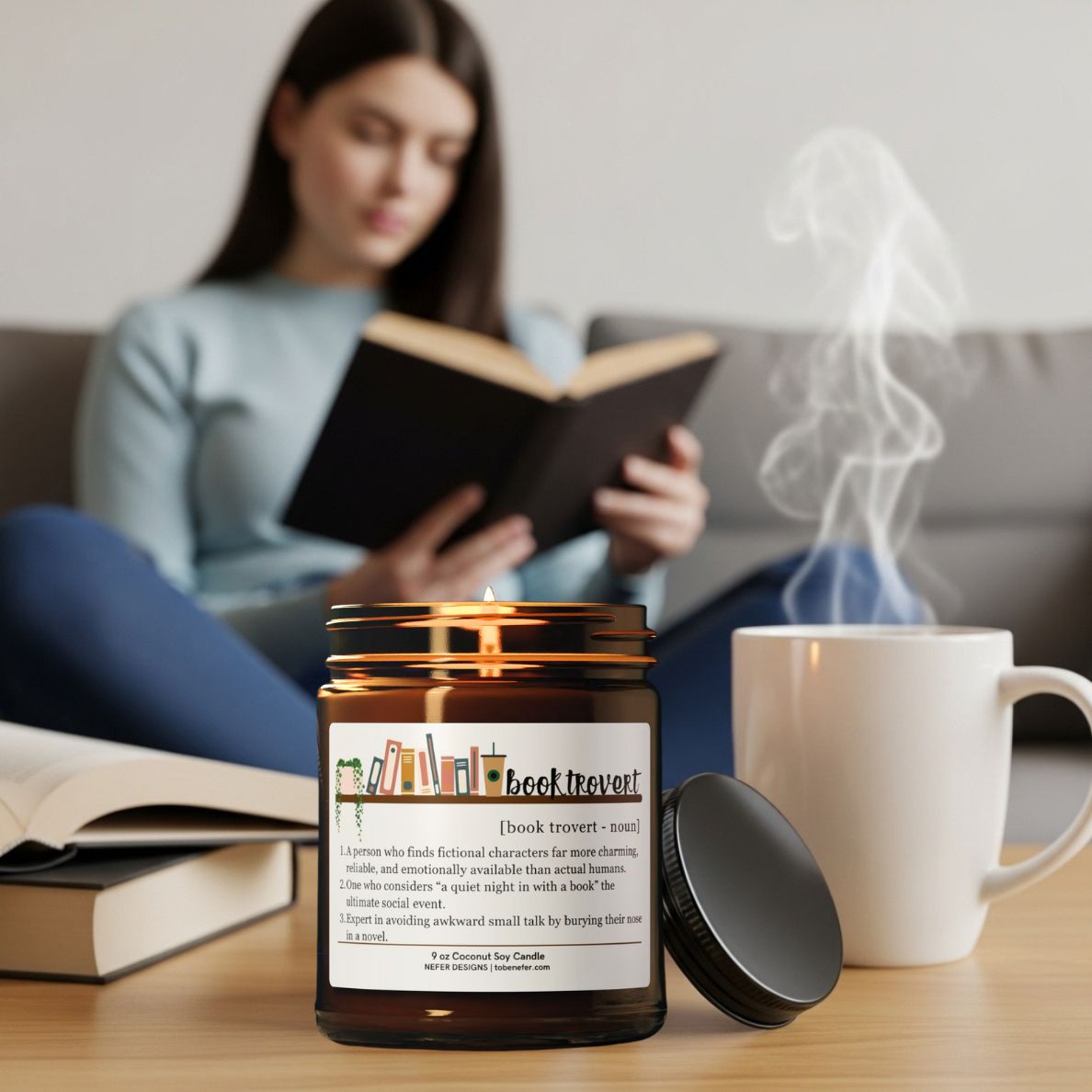 Woman reading a book with a lit  book themed candle and steaming mug on a table in the foreground.