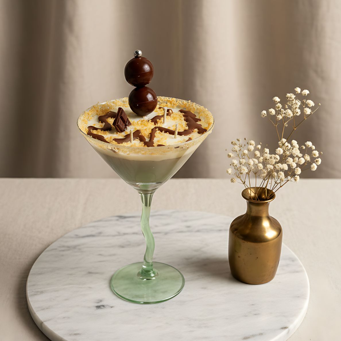 uxury Chocolate Martini candle in a green wavy-stem martini glass on a round marble tray, next to a small gold vase with baby's breath flowers. The candle's chocolate drizzle and embeds are clearly visible, set against a soft, neutral background.