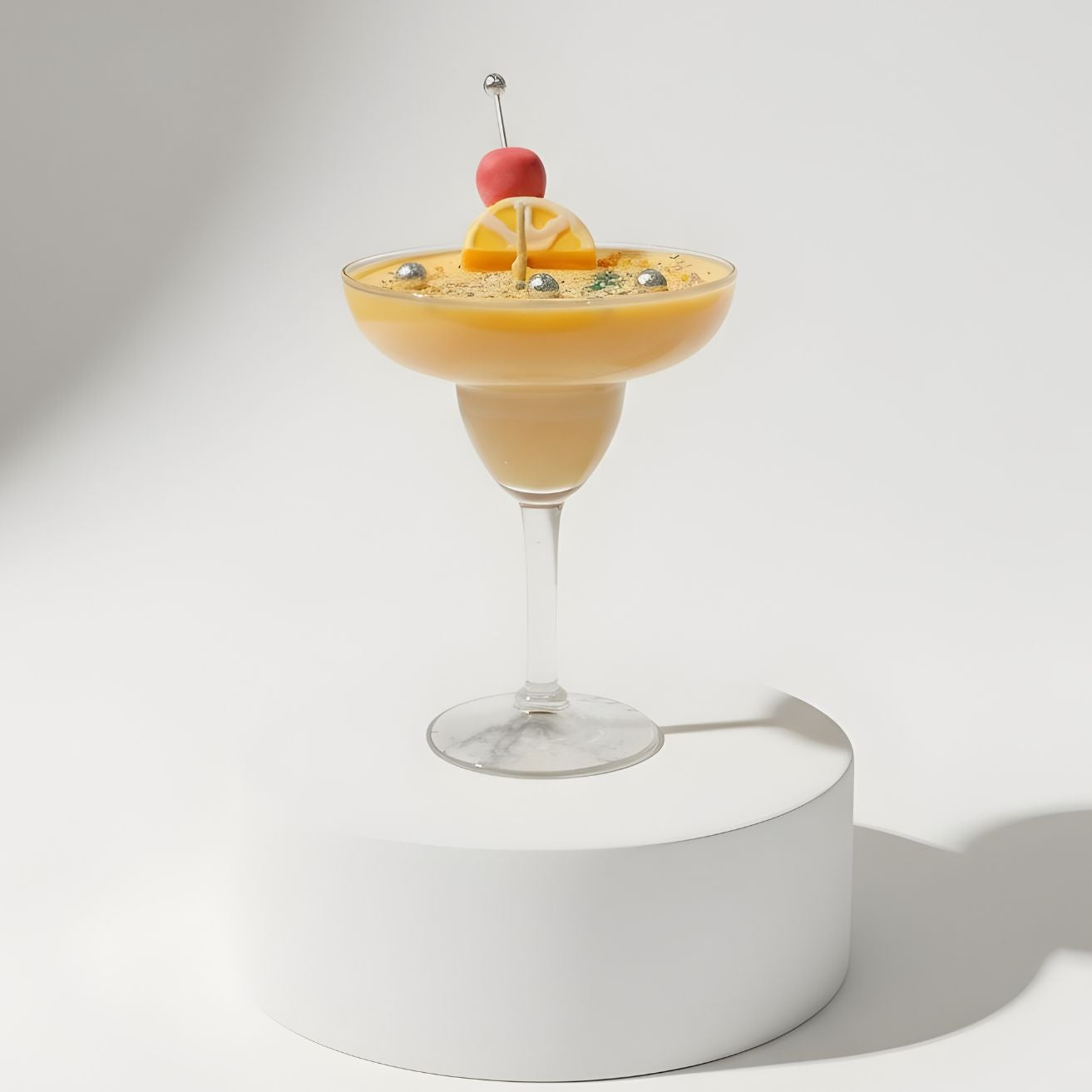 A yellow cocktail candle in a margarita glass, garnished with a pink and yellow candy skewer, displayed on a square white pedestal in a minimalist, bright setting.
