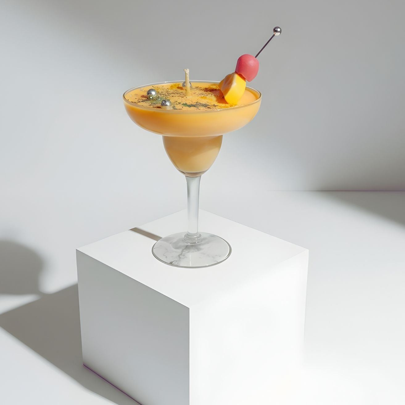 A yellow cocktail candle in a margarita glass, garnished with a cherry and lemon wedge, displayed on a short, round white pedestal in a brightly lit studio.