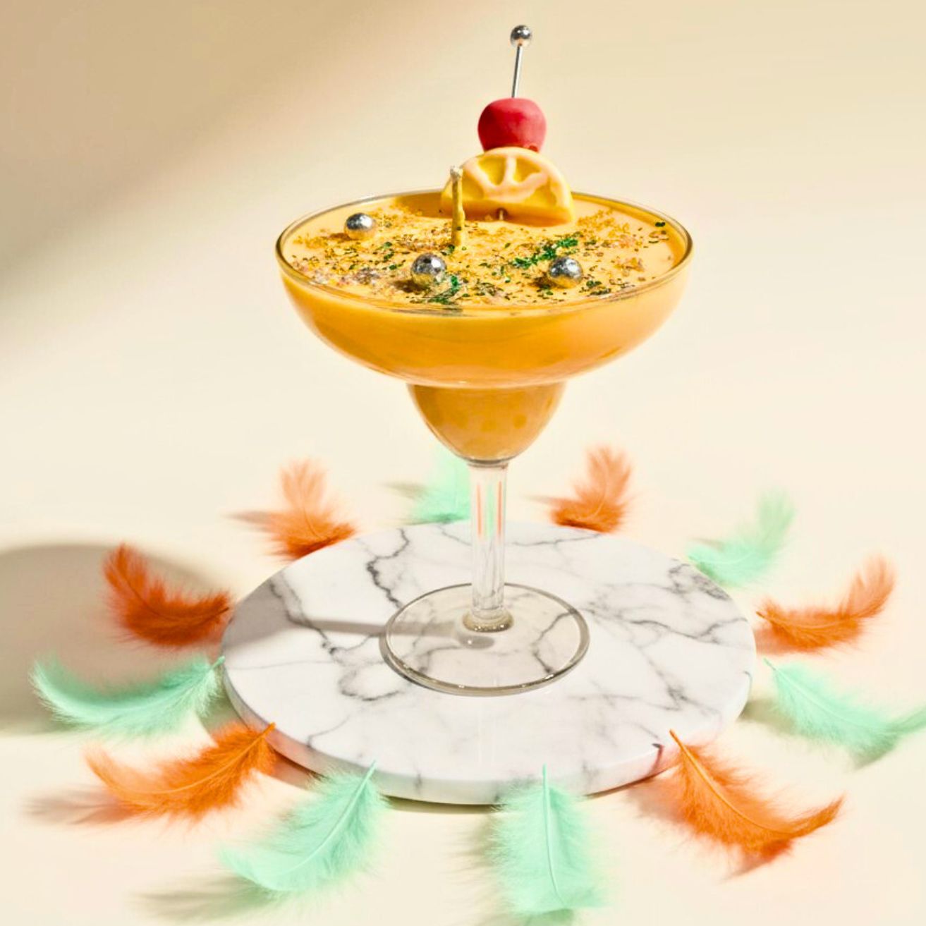 A yellow cocktail candle in a margarita glass, sitting on a white marble coaster surrounded by orange and mint green feathers, against a beige background.