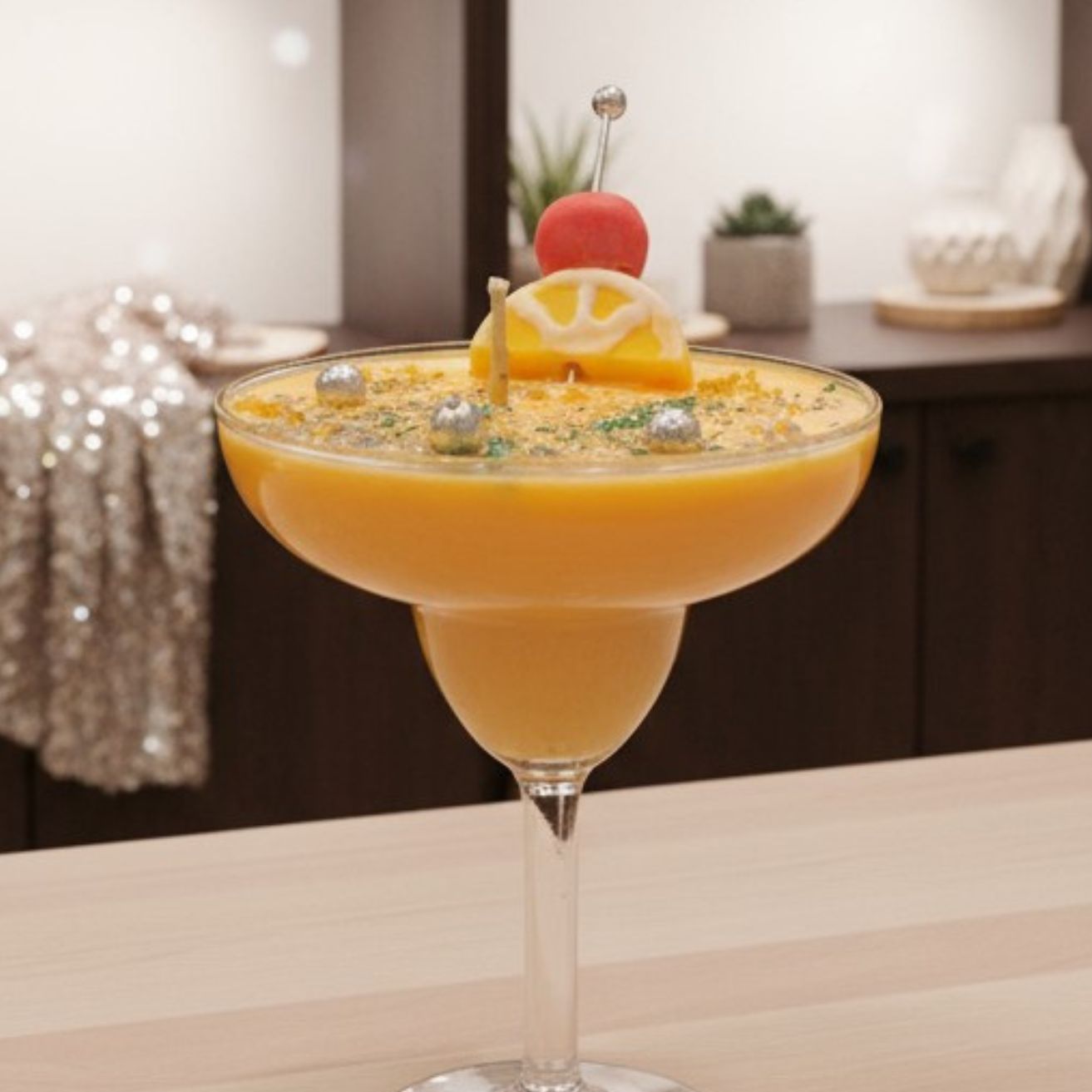 A yellow cocktail candle in a margarita glass, placed on a wooden counter in an indoor setting with dark cabinets and a silver, sequined cloth draped in the background.