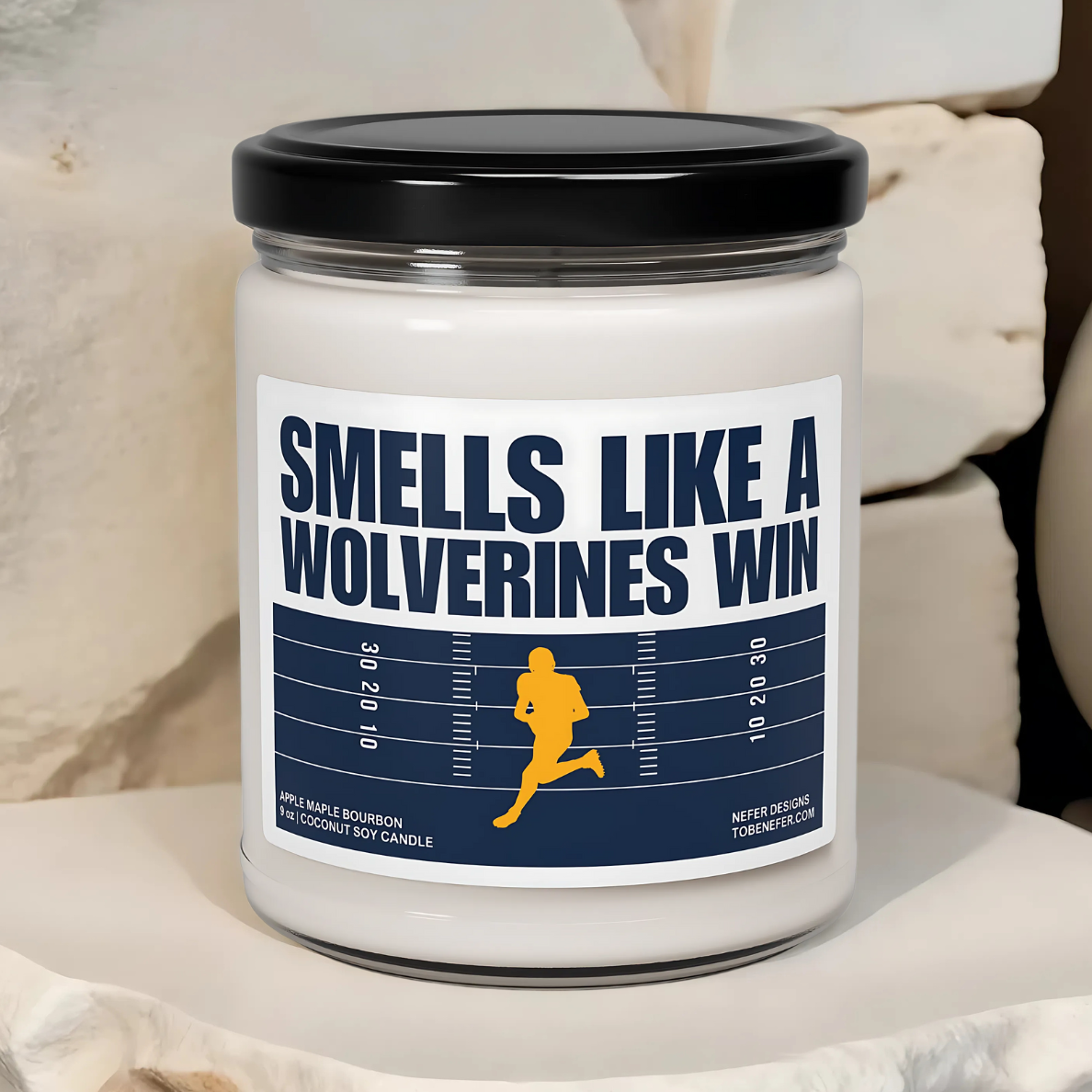A Michigan Wolverines-themed soy candle with a label that says 'Smells Like a Wolverines Win | Lucky Game Day,' sitting on a wooden surface.