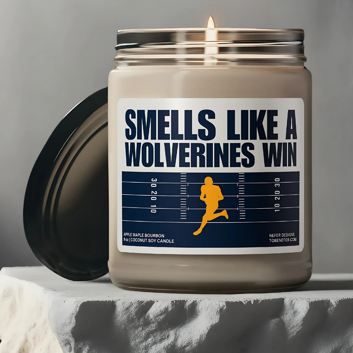 A Michigan Wolverines-themed soy candle with a label that says 'Smells Like a Wolverines Win | Lucky Game Day,' sitting on a wooden surface.