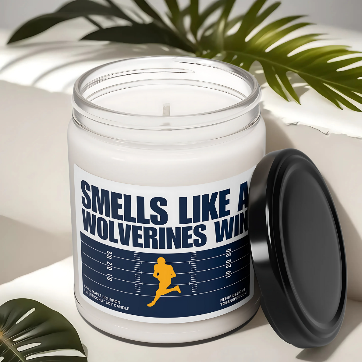 A Michigan Wolverines-themed soy candle with a label that says 'Smells Like a Wolverines Win | Lucky Game Day,' sitting on a wooden surface.
