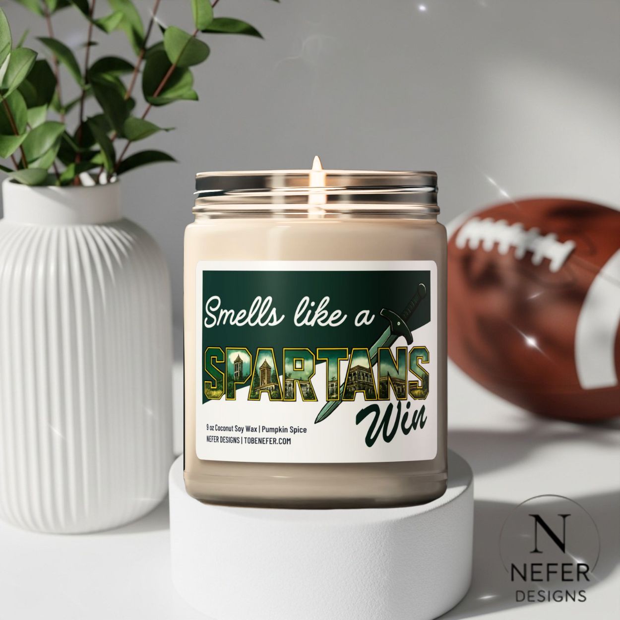 Candle with a label reading 'Smells like a Spartans Win' on a white surface with a football and plant in the background.