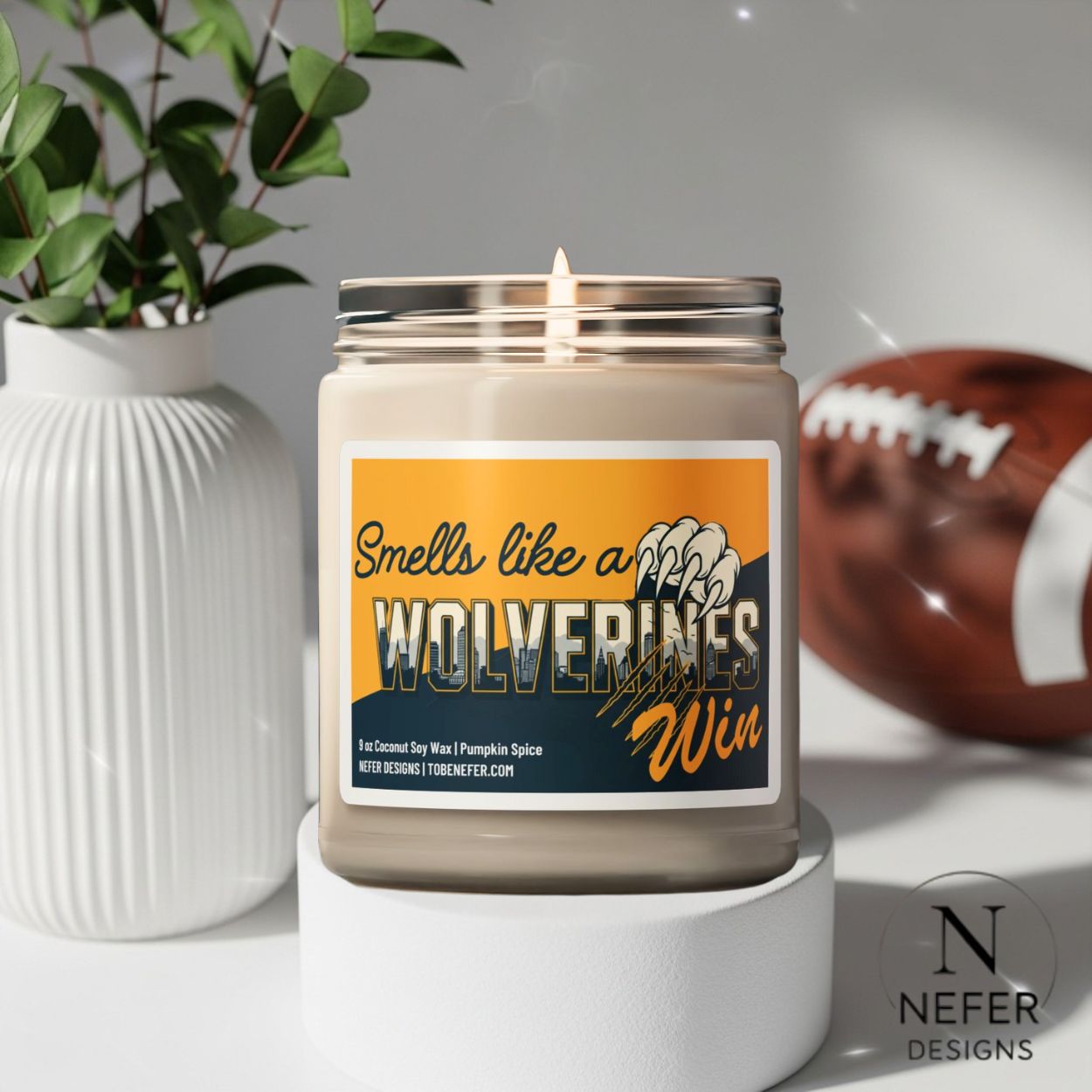 Candle with a label reading 'Smells like a Wolverines Win' on a white surface with a football and plant in the background.