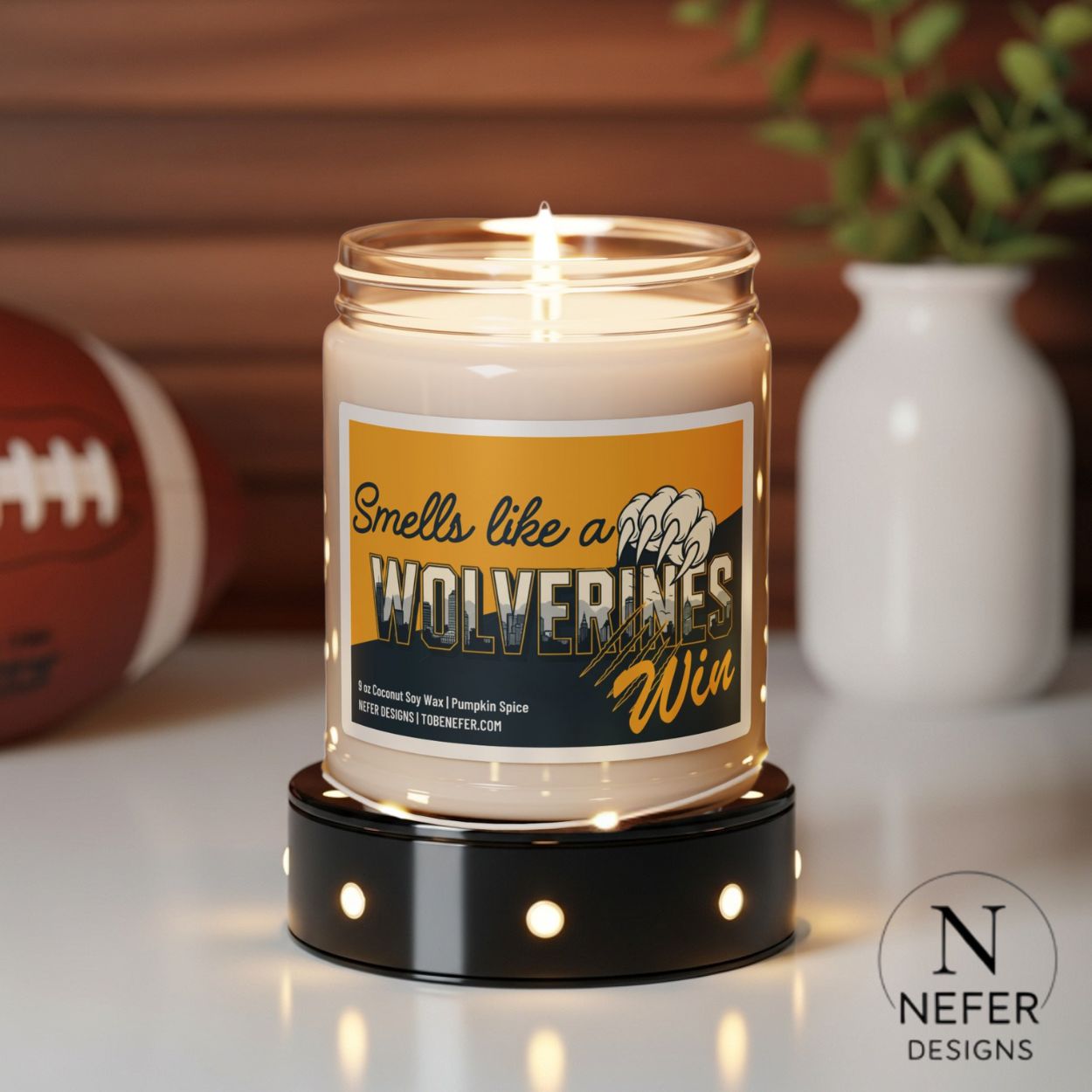 Candle in a jar with a label reading 'Smells like a Wolverines Win' on a decorative stand, with sports ball and vase in the background.