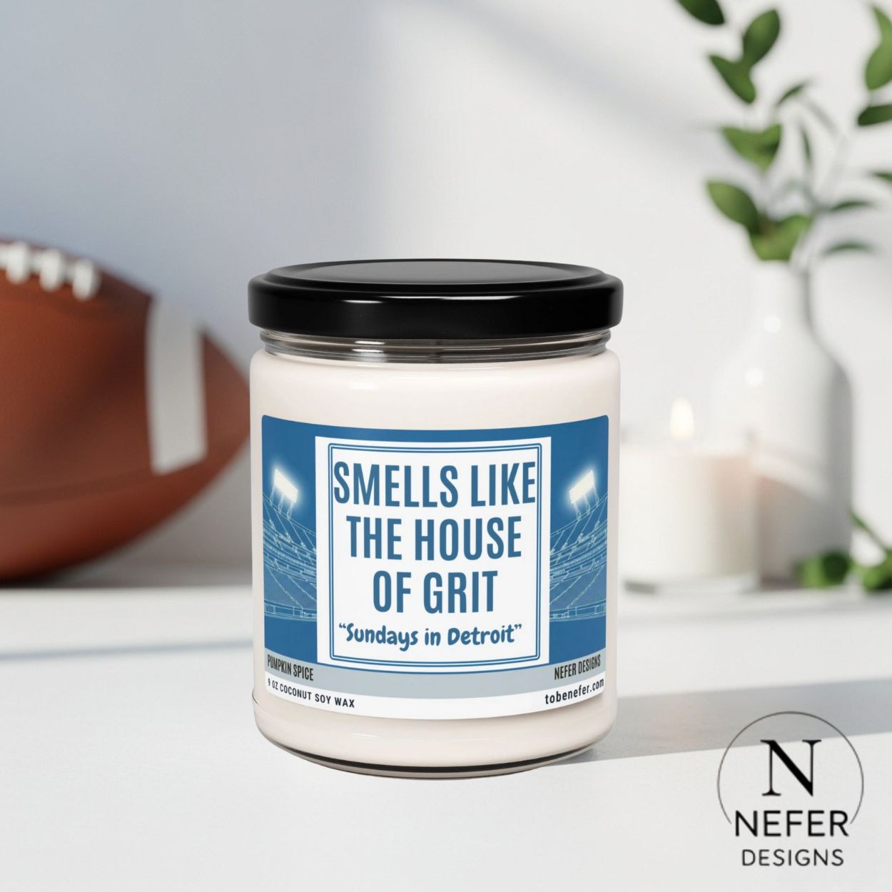 Candle with a label reading 'Smells Like the House of Grit' by Nefer Designs on a surface with a football in the background.