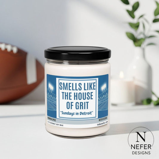 Candle with a label reading 'Smells Like the House of Grit' by Nefer Designs on a surface with a football in the background.